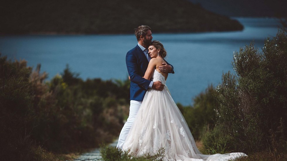 A post-wedding embrace from a bride and groom on the hill above Punga Cove with scenic background views of Endeavour Inlet in the Marlborough Sounds of New Zealand