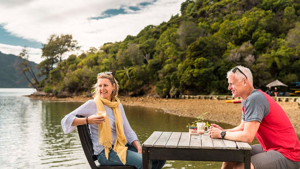 A relaxed couple drink and enjoy Boatshed Cafe and Bar coffee on the jetty at Punga Cove in the Marlborough Sounds in New Zealand