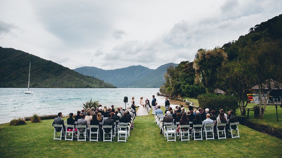 An outdoor wedding ceremony is held on the front lawn at Punga Cove with scenic background views of Endeavour Inlet in New Zealand's South Island Marlborough Sounds