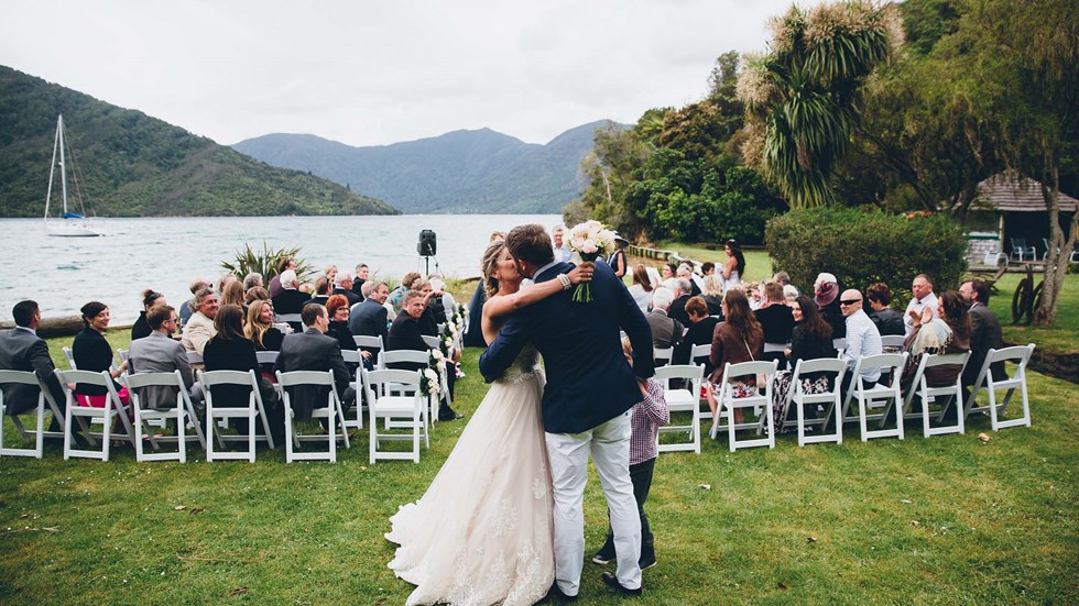 A newly married bride and groom kiss on the front lawn of Punga Cove following the ceremony with watching guests in the Marlborough Sounds of New Zealand