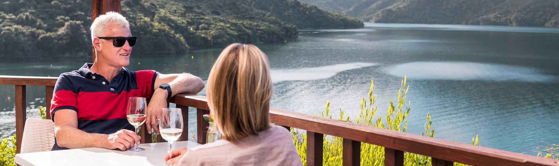 A couple enjoying their glasses of wine from the Punga Fern Restaurant located at Punga Cove with sweeping views of the Endeavour Inlet in New Zealand's top of the South Island Marlborough Sounds.