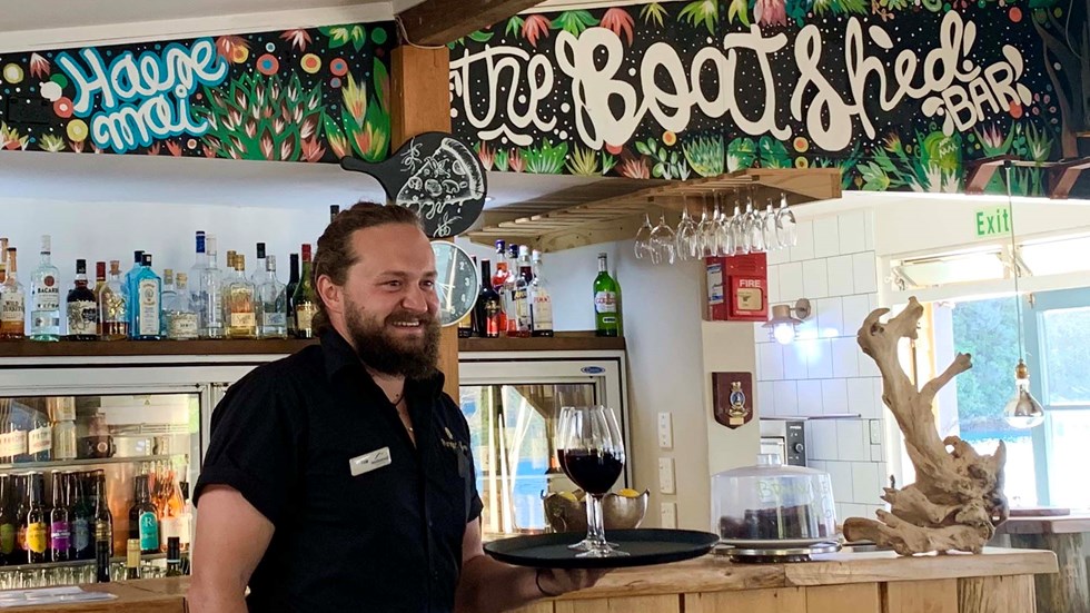 A friendly bartender from the Boatshed Cafe and Bar delivers glasses of Pinot Noir wine to guests at Punga Cove located in the Marlborough Sounds of New Zealand's South Island