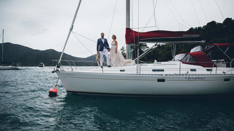 A Bride and Groom hold hands and stand on a moored yacht in the Punga Cove bay with Endeavour Inlet background views in the Marlborough Sounds of New Zealand