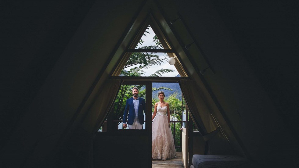 A bride and groom stand holding hands at the entrance of a Koru Chalet room at Punga Cove with a scenic lush Punga fern forest behind them in the Marlborough Sounds, New Zealand