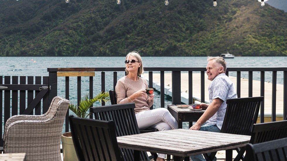 A couple sit at the Boatshed Cafe and Bar outdoor seating area with coffee enjoying the scenic views of the Endeavour Inlet in the Marlborough Sounds, New Zealand
