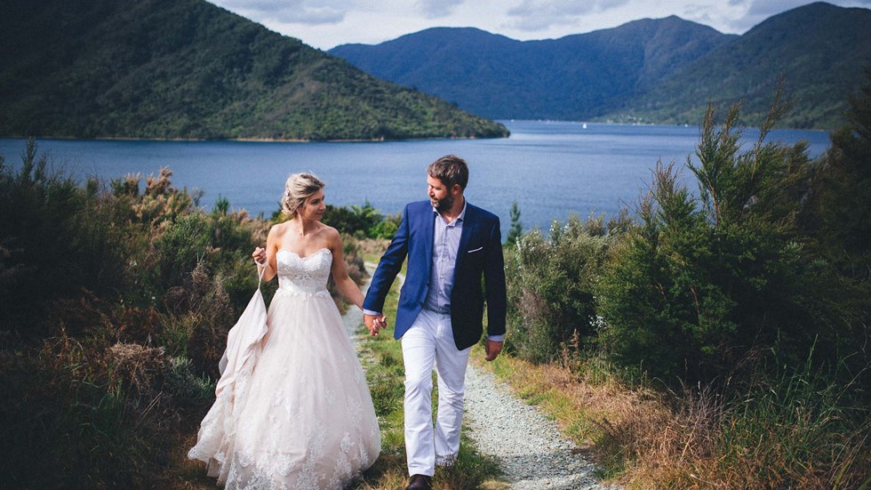 A Bride and Groom walk along a hill above Punga Cove with scenic views of Endeavour Inlet in the background in the Marlborough Sounds of New Zealand